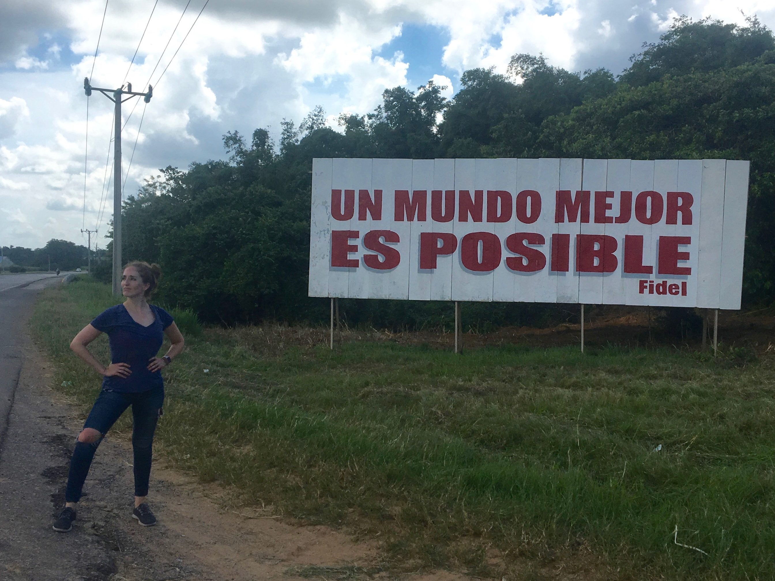 Dr. Michaelanne Thomas stands by a billboard sign on the side of a gravel road that reads, 'Un mundo mejor es posible, Fidel'