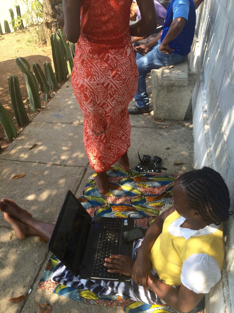 A woman sits on the ground leaning against a wall with an open laptop in her lap. Nearby, people are on their phones, browsing social media.